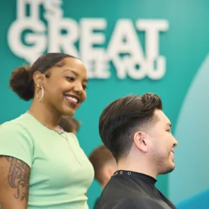 A female stylist stands behind a male client in a hairdresser cape